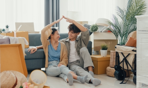 Happy couple sit on floor in front of sofa couch smile looking at camera making house sign in living room at new house